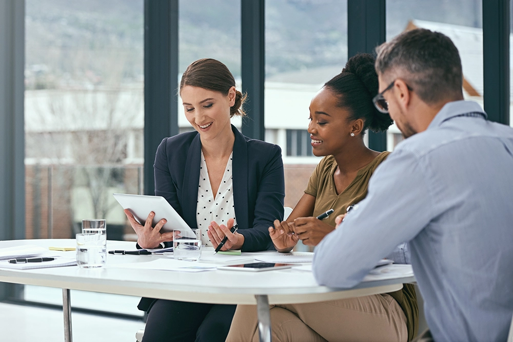 Three colleagues meeting around a table and reviewing work on a tablet.